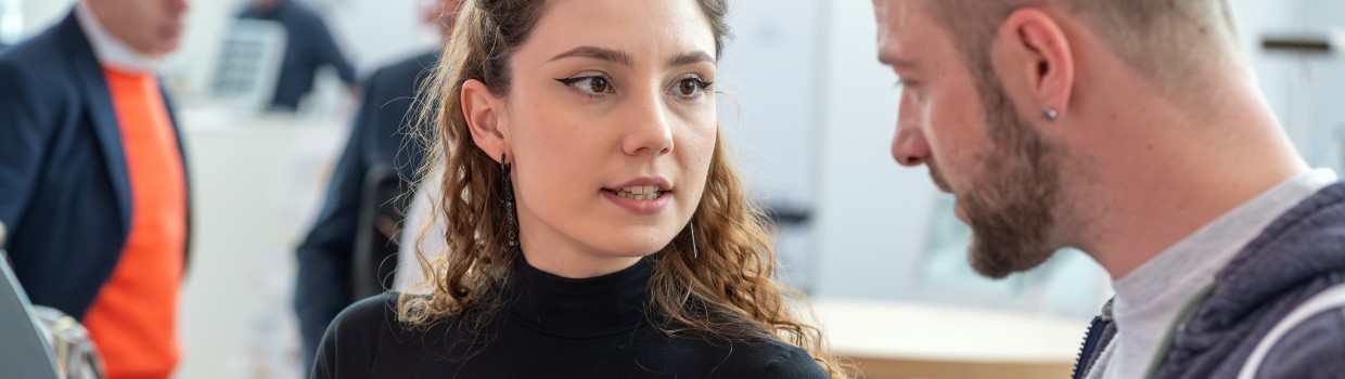 A woman with long curly hair wearing a black turtleneck shows a man with a beard a dental model at the zahntechnikplus