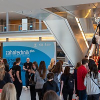 Menschenmenge im Eingangsbereich der zahntechnikplus vor einer blauen Begrüßungswand; einige Personen stehen an, andere fahren auf einer Rolltreppe in die obere Etage des modernen Messegebäudes.