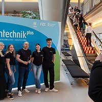Fünf Personen posieren vor einer blauen Fotowand mit dem Logo der zahntechnikplus im Messegebäude; rechts fotografiert eine Frau die Gruppe, im Hintergrund fahren Menschen die Rolltreppe hinauf.