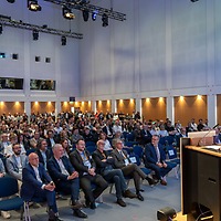 Ein Redner steht am Pult vor einem voll besetzten Vortragssaal der zahntechnikplus; das Publikum blickt zur Bühne, der Raum ist hoch und modern mit Bühnenbeleuchtung an der Decke.