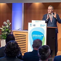 Ein Mann im Anzug spricht gestikulierend am Rednerpult mit zahntechnikplus-Logo vor einem sitzenden Publikum; links neben dem Pult steht ein Blumenarrangement, Hintergrund aus Holzpaneelen.