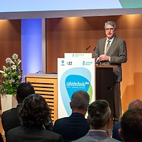 Ein Mann im gemusterten Sakko spricht bei der zahntechnikplus am Rednerpult mit Veranstaltungslogo vor einem sitzenden Publikum; links daneben steht ein Blumenarrangement, im Hintergrund Holzverkleidung.