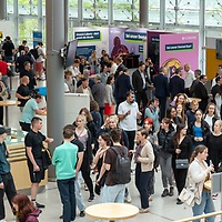 Zahlreiche Besucherinnen und Besucher bewegen sich durch die helle Messehalle der zahntechnikplus mit mehreren bunten Ausstellerständen und großen Fenstern im Hintergrund.