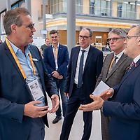 Fünf Männer in Anzügen unterhalten sich im Foyer der zahntechnikplus; einer trägt ein Namensschild mit orangefarbenem Band, im Hintergrund sind weitere Teilnehmende und moderne Architektur sichtbar.
