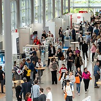 Zahlreiche Besucherinnen und Besucher bewegen sich durch die helle Ausstellungshalle der zahntechnikplus mit vielen Messeständen, Vitrinen und Informationsdisplays entlang breiter Gänge.