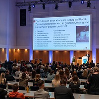 Vortragssaal der zahntechnikplus mit sitzendem Publikum an Tischen; auf der Bühne steht ein Redner neben einer großen Leinwand mit einer Präsentationsfolie