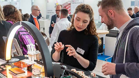 Eine Frau erklärt einem Mann am Messestand der zahntechnikplus ein Zahnmodell unter einer Bogenlampe; auf dem Tisch liegen weitere Dentalmodelle, im Hintergrund stehen weitere Besucherinnen und Besucher.