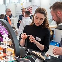 Eine Frau erklärt einem Mann am Messestand der zahntechnikplus ein Zahnmodell unter einer Bogenlampe; auf dem Tisch liegen weitere Dentalmodelle, im Hintergrund stehen weitere Besucherinnen und Besucher.