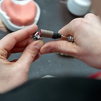 Zwei Hände bearbeiten bei der zahntechnikplus mit einem rotierenden Werkzeug ein kleines Zahntechnik-Bauteil; im Hintergrund liegt ein rosa Zahnfleischmodell.