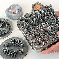 Eine Person hält bei der zahntechnikplus eine Metallplatte mit zahlreichen gestützten 3D-gedruckten Zahntechnik-Elementen; auf dem Tisch liegen weitere Bauplattformen mit dentalen Strukturen.