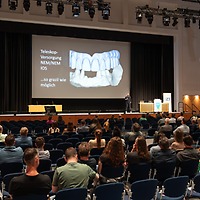 Ein Vortragssaal bei der zahntechnikplus mit Publikum in Reihen bestuhlter Sitze; auf der Bühne steht ein Redner neben einer großen Projektion eines digitalen Zahnmodells