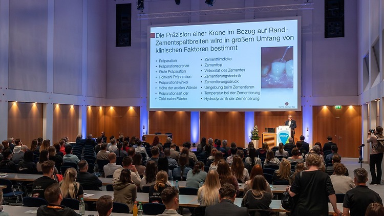 Ein voll besetzter Vortragssaal bei der zahntechnikplus, in dem ein Redner auf der Bühne eine Präsentation über Kronenpräzision zeigt; auf der Leinwand sind Textinformationen und ein Zahnfoto zu sehen.